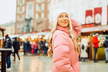 A cute blonde in a hat and a pink jacket walks around the city, winter fairs. Christmas mood smile