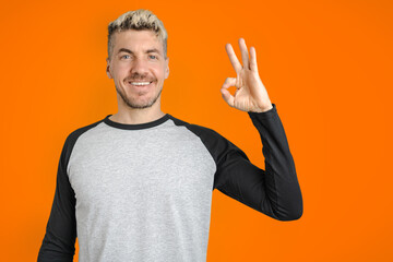 Young Caucasian man doing Ok gesture with finger and smiling. Isolated orange background