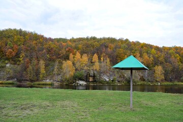Fototapeta premium Metal beach umbrella on a green lawn against the background of a river, mountain and waterfall on an autumn day 