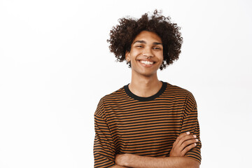 Close up of handsome modern hispanic guy with afro hair, smiling happy, cross arms on chest and...