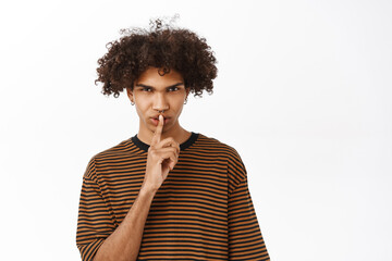 Fototapeta na wymiar Keep quiet. Serious hispanic boy shushing, say shh, showing taboo gesture with finger pressed to his mouth, standing over white studio background