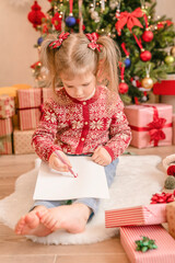 Child girl writing letter to Santa Claus at home while sitting on the floor
near Christmas tree. Merry Christmas! Adorable child writing letter to Santa Claus