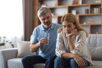 Worrying middle aged european man gives glass of water and calms depressed woman on sofa in living room