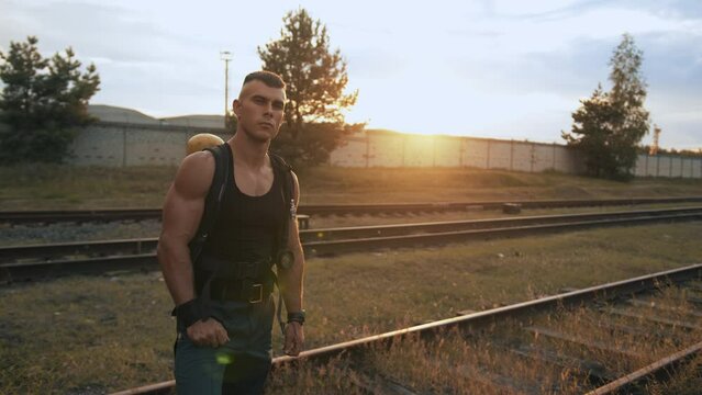 Young muscular man with balloon drager on his back stands on rails at marshalling yard at sunset and looks at his watch. Slow motion