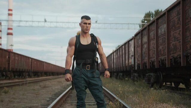 Bodybuilder in a black sleeveless t-shirt resting between exercises in training at railway yard and looking at the sky