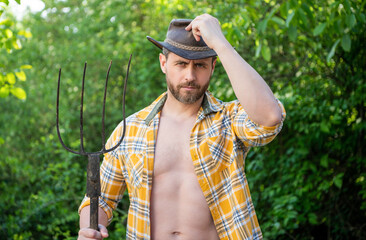 Serious cowboy man tipping cowboy hat holding pitchfork. Cowboy in garden natural background
