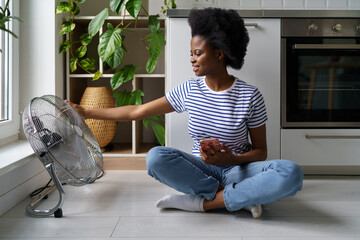 Satisfied African American woman enjoying cool air coming from behind electric fan and relaxing...