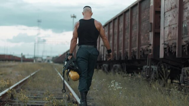 Bodybuilder goes along the rail keeping the balance past freight train and carries balloon dragger in his hands. Back view