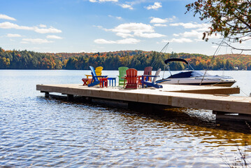 Fototapeta premium Motorboat tied up to a wooden pier on a lake on a sunny autumn day. Colourful armchairs are on the pier.