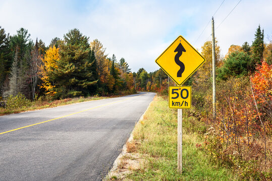 Wanrning road sign along with a speed limit sign on a winding forest road on a sunny autumn day