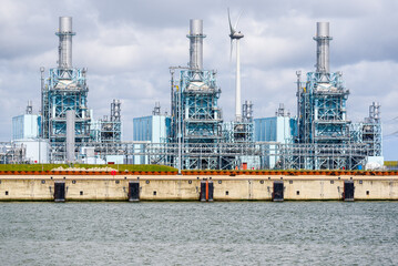 Fototapeta premium Gas fired power station on a harbor on a cloudy summer day. A wind turbine is in background. Eemshaven, Netherlands.