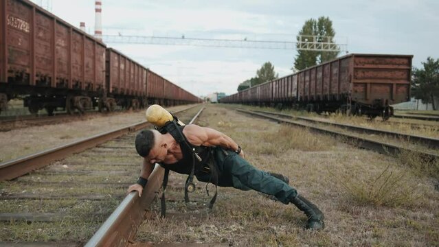 Bodybuilder with balloon dragger on the back is training at railway yard and doing push-ups from the rails on one arm