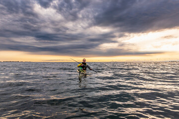 A sea kayaker (wearing expedition safety gear) sits in his boat with the sun setting behind him on Georgian Bay.