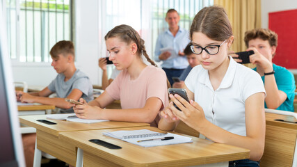 Young students with smartphones sitting in class room. Teacher explaining something to them.