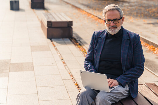 Remote Working Concept. Adult Caucasian Gray-haired Bearded Businessman In Formal Clothes Benefiting By Beautiful Sunny Autumn Weather And Working Outside With His Laptop. Looking At Camera. High