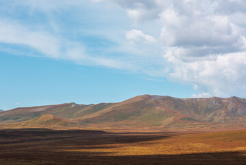 Motley autumn landscape with sunlit high mountain plateau and mountain range under dramatic cloudy sky. Vivid autumn colors in mountains. Sunlight and beautiful shadows of clouds in changeable weather