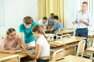 Fototapeta na wymiar Interested teenage students divided into small groups, trying to solve teacher assignments together in classroom, using mobile phones