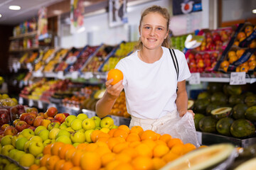 Portrait of teenage blonde girl customer buying sweet oranges at grocery shop