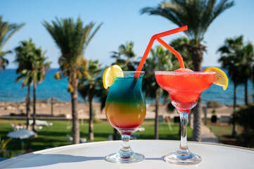 Colourful cold Rainbow Paradise and Strawberry daiquiri cocktail drinks served in glasses at pool bar overlooking blue pool, sea and palm trees, relax and holidays at sea