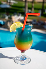 Colourful cold Rainbow Paradise cocktail drink served in glass at pool bar overlooking blue pool, sea and palm trees, relax and holidays at sea