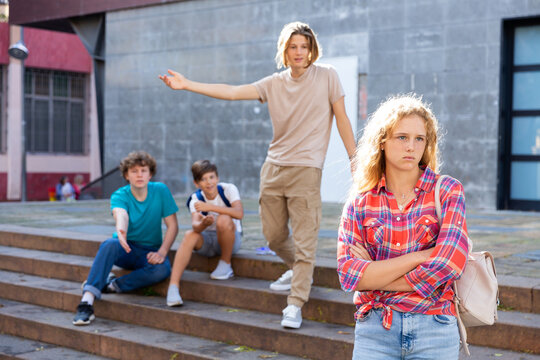Sad young girl with backpack standing near school building. She's offended by her friends who trying to talk with her in background.