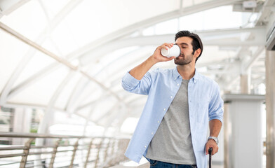 The man drinks a cup of coffee and pulls the luggage while he is waiting for his friend at the airport.
