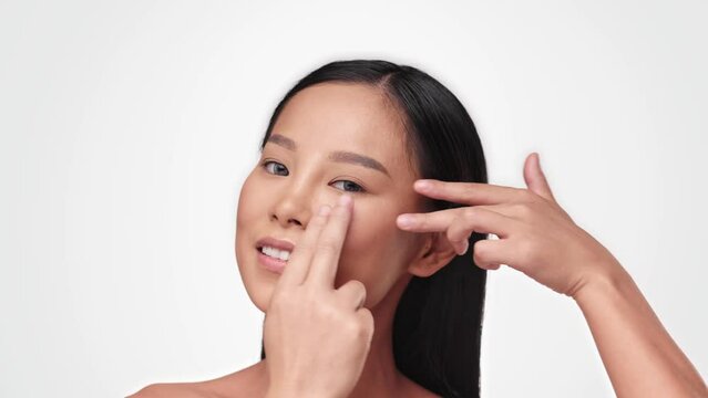 Portrait of pretty millennial woman touching her face while looking to camera. Asian Female model with naked shoulders and nude make up massaging her face after applying cream. Beauty treatment.