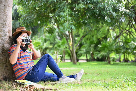 A middle-aged woman sat comfortably on the green lawn under a large tree. Bring a digital camera to capture beauty of garden. Tourists wear glasses and brown straw hats, bright shirt holding a camera
