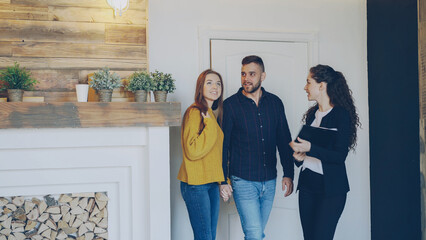 Cheerful female realtor is meeting beautiful young couple in new house opening door showing documents and talking to clients. Happy people and selling real estate concept. - obrazy, fototapety, plakaty