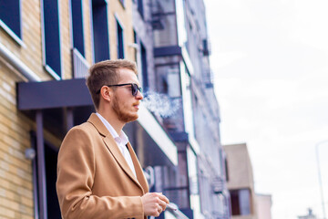 A young guy in sunglasses and a beige stylish coat smokes on the street in the city. Bad habit