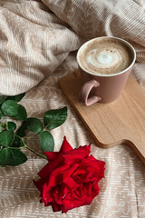 A red rose and a pink coffee mug are on the bed with beige linens. Surprise for the morning. Festive breakfast