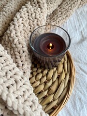 A black lighted candle in glass on a rattan stand and a plush beige plaid on the bed in the room. Selective focus. Create a cozy atmosphere by adding candles to your home decor. Flatlay