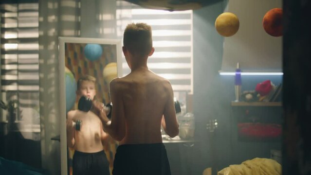 A focused school boy do dumbbells curls stands in front of the mirror in his bedroom. Nude upper body and black shorts. Prepares for a match. Fitness, weight traning concept.