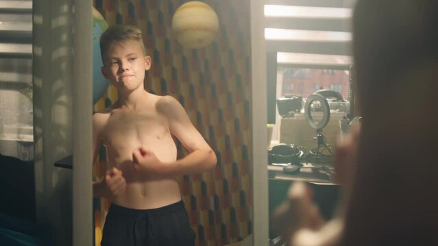 Muscular schoolboy examines his body after weight training exercise in front of mirror in his room. Muscular abs and shoulders. Bare upper body. Weight training, athletes, fitness, physical activity.
