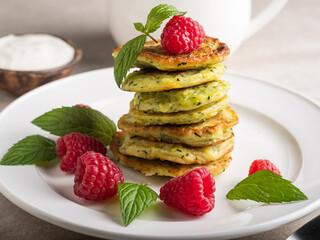 Stack of fresh homemade zucchini pancakes topped with raspberries and sour cream. Green tea in a white cup.