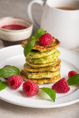 Stack of fresh homemade zucchini pancakes topped with raspberries and sour cream. Green tea in a white cup.