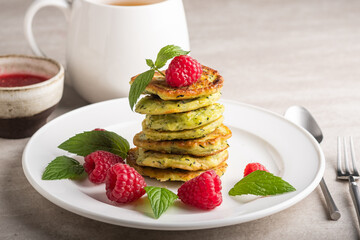 Stack of fresh homemade zucchini pancakes topped with raspberries and sour cream. Green tea in a white cup.