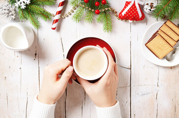 Female hand holding a cup of cappuccino with cookies on wooden background with Christmas thematic decorative elements. Copy space, top view.