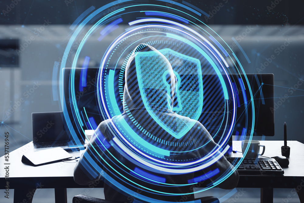 Poster hacker at workplace using laptop with glowing shield hologram on blurry office interior background. 