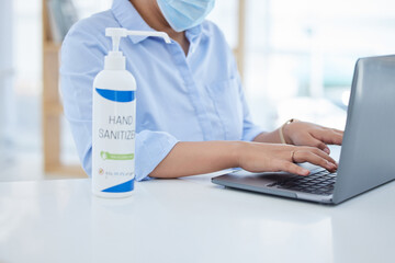 Covid, hand sanitizer and a woman in office typing on laptop with face mask. Online work, hygiene and businesswoman with alcohol spray on desk. Virus prevention, safety and cleaning hands in pandemic