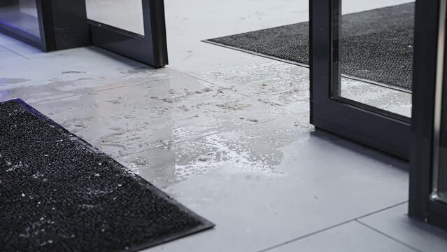 Snow and slush on wet and slippery tiles floor surface. People in snow-covered shoes enter and go out store. Fleecy rugs mats lies near sliding automatic doors at entrance to building shopping center