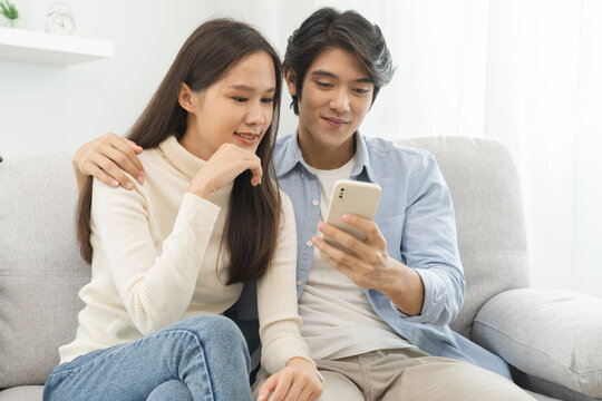 Happy couple love at home,beautiful two asian young spending good time together,bonding to each other and smiling romantic on sofa in living room while man embrace woman using smartphone, mobile phone