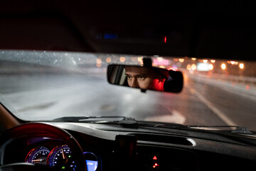 Look of man is reflected in rear-view mirror traveling in car in evening hurries home after work. Speedometer with neon lights and eyes young guy working as driver or cabbie and enjoying city at night