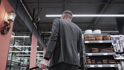 Male buyer with plastic basket walks through grocery store and looks around in search of goods. Customer 30 35 years old in jacket in shopping mall centre supermarket. Sales promotions discounts