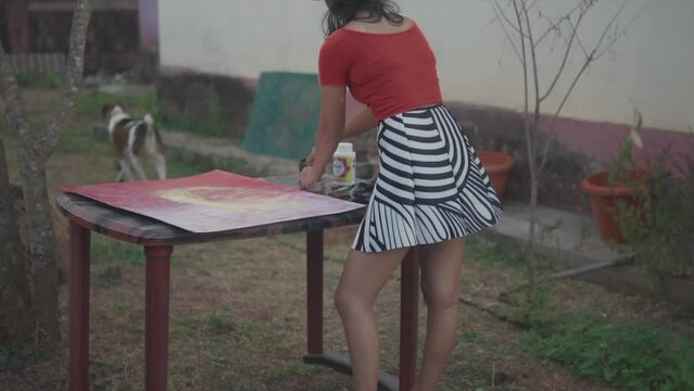 Slow panning shot of an indian artist dressed in red top, striped skirt and sporty running shoes painting a picture with acrylic paints and a jack russell dog running through the garden in slow motion