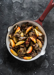 Fried chicken liver with onions and apples in a ceramic frying pan on a dark background, top view