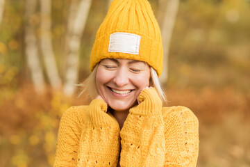 Close-up portrait of a cheerful blonde woman with closed eyes in a yellow hat in an autumn park , on a blurry background.