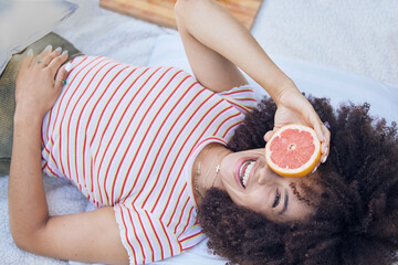 Fruit, picnic and summer with a happy black woman holding a grapefruit while lying on a blanket outdoor. Food, health and wellness with a female laughing or joking while outside to relax from above