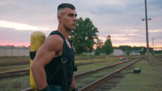 Portrait of handsome muscular man with bottle drager behind his back on the railroad tracks in the summer at sunset. Slow motion