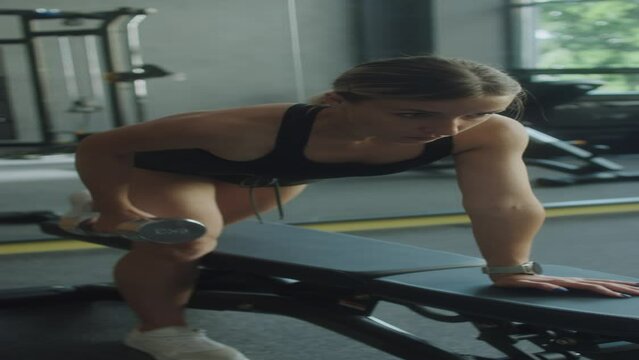 Vertical shot of woman in sportswear doing single arm dumbbell row exercise on gym bench